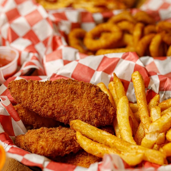 Crispy chicken tenders and golden french fries in a red and white checkered basket, with onion rings in the background.