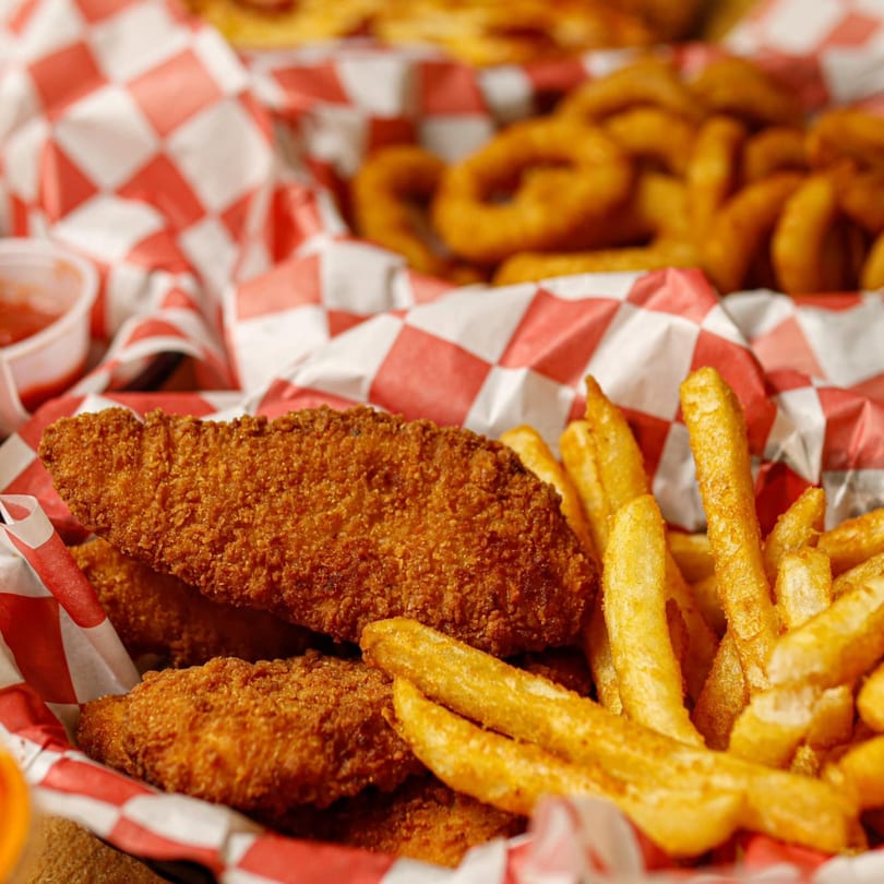 Crispy chicken tenders and golden french fries in a red and white checkered basket, with onion rings in the background.