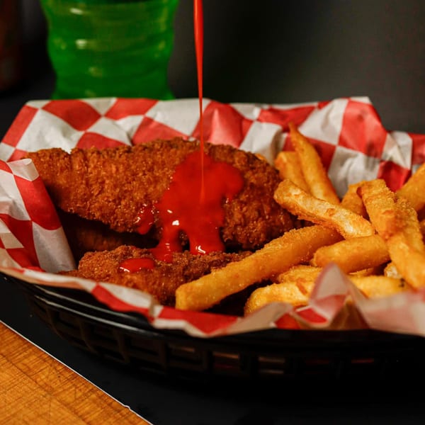 Crispy chicken tenders with fries in a basket, red sauce pouring over the tenders.